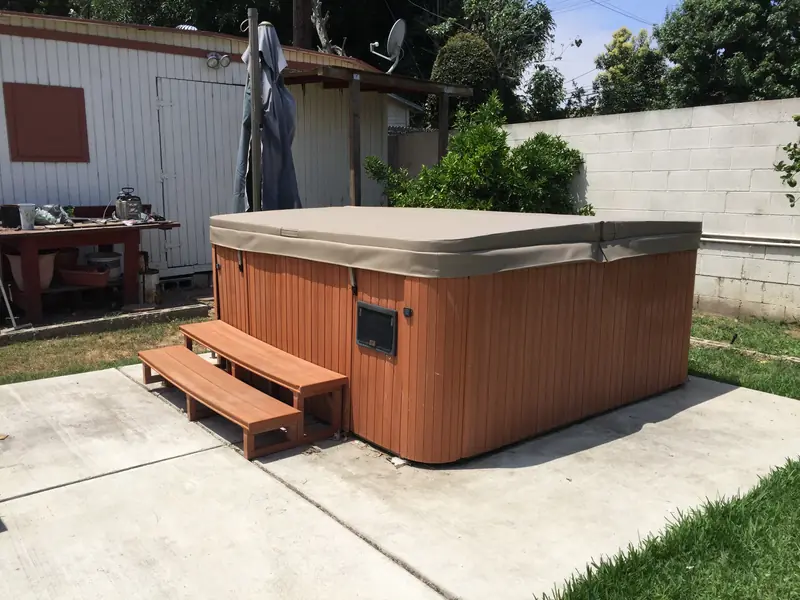 A rectangular tan two-panel spa cover on a freestanding spa with a red-stained timber cabinet and timber steps, standing on a concrete slab in a lawned backyard beside a block wall.