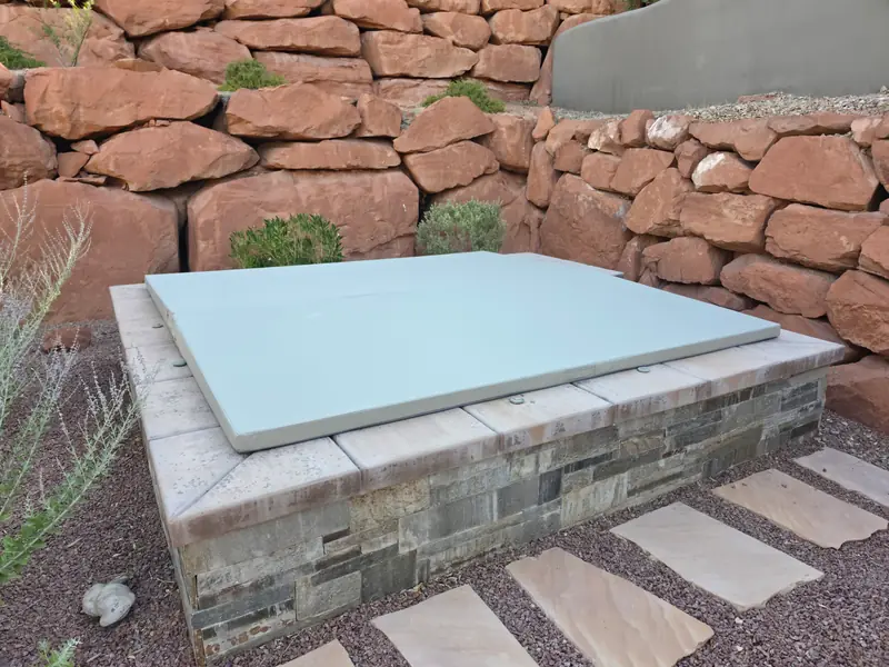 A square pale-grey spa cover with a single centre fold, sitting flush on an in-ground spa built into a stone-faced surround, with a wall of large red boulders and dry planting behind it.