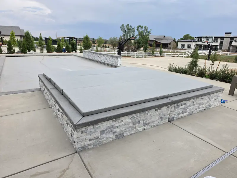 A rectangular light-grey spa cover on a raised in-ground spa faced in stacked grey stone, set into a concrete pool deck, with a swimming pool alongside and houses beyond a white fence.