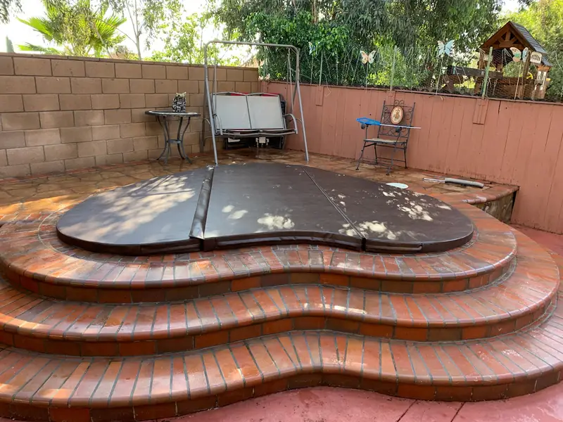 A kidney-shaped dark-brown spa cover following the curve of an in-ground spa raised on three tiers of curved brick steps, in a walled patio with a swing seat and bistro chairs behind.