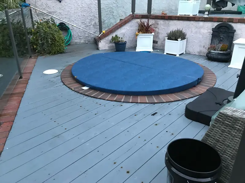 A round mid-blue spa cover on an in-ground spa ringed with brick and set into a grey painted timber deck, with white planters and a wall fountain along the back wall.
