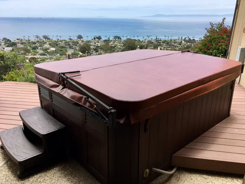 A rectangular dark red-brown spa cover with rounded corners on a freestanding spa with a dark cabinet, standing on a red-stained timber deck; below and behind, a hillside town, palm trees and the open ocean.