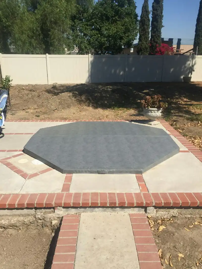 An eight-sided grey spa cover on an in-ground spa set flush into a concrete patio edged with red brick, with a white fence, tall narrow conifers and bare planting beds behind.