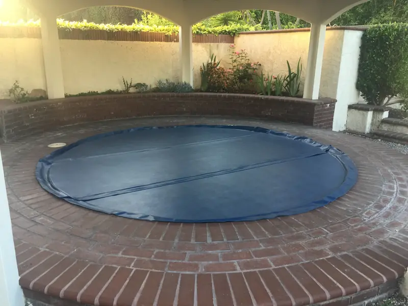 A large round navy-blue spa cover lying on a circular in-ground spa set into a brick patio, beneath a white arched shelter with planting behind a low brick wall.