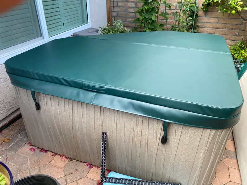 A rectangular dark green spa cover with cut corners on a freestanding spa with a mottled beige cabinet, tucked between a house wall with green shutters and a brick planter.