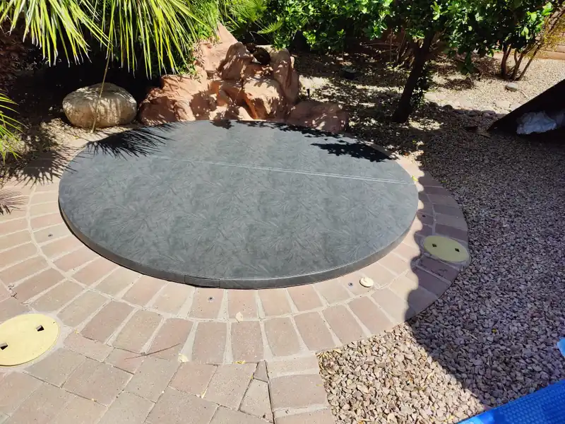 A round grey-green spa cover on an in-ground spa set into a circle of brick pavers, edged by red rock, gravel and a palm, with dappled shade across the cover.
