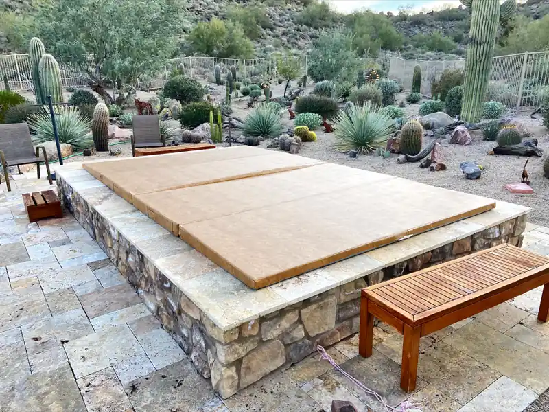 A large rectangular tan spa cover in three folded panels on an in-ground spa raised in a stone-faced plinth, with a pale stone patio in front and a desert garden of tall columnar and barrel-shaped cactus behind.