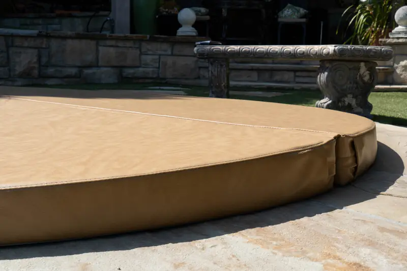 A close view along the edge of a round tan spa cover, showing the stitched seam across the centre, the folded corner flap and the depth of the padded skirt, with a carved bench behind.