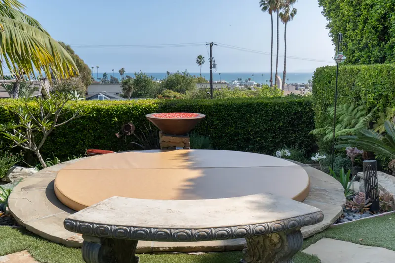 The same round tan spa cover seen from further back, the stone terrace ringed by planting, the fire bowl of red chippings behind it and the coastline and ocean visible over the hedge.