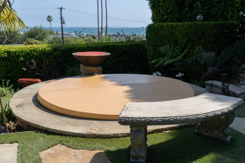 A round tan spa cover on a circular stone-paved terrace, with a fire bowl of red chippings on a stone plinth behind it, a curved carved bench in the foreground, and palms and the ocean beyond a clipped hedge.