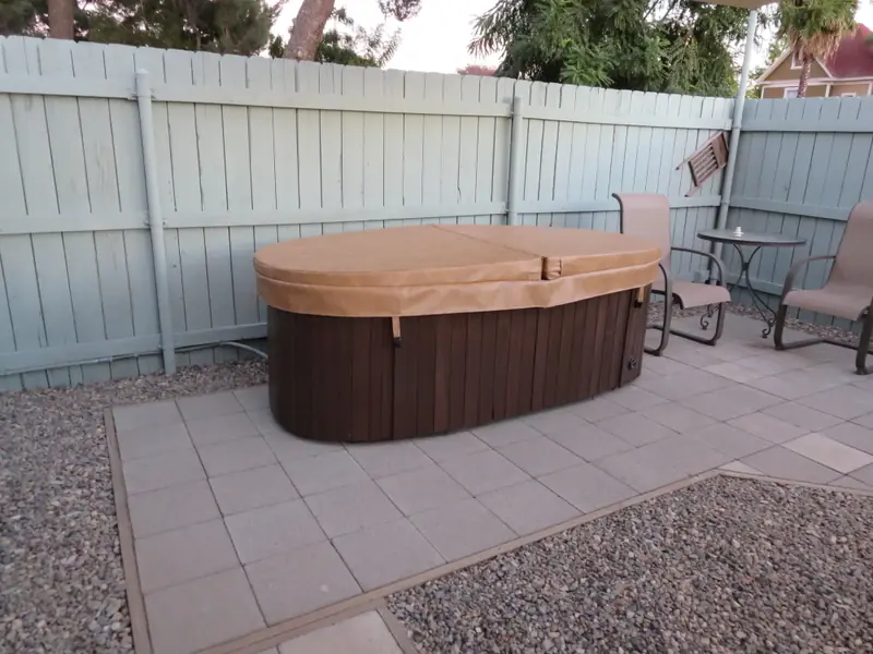 An oval tan spa cover on a freestanding spa with a dark stained timber cabinet, on a paved courtyard against a pale green board fence, with patio chairs and a bistro table alongside.