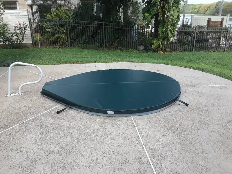 A pear-shaped dark green spa cover, rounded at one end and tapering to a point at the other, on an in-ground spa flush with a concrete deck, with a handrail alongside and open water beyond the fence.