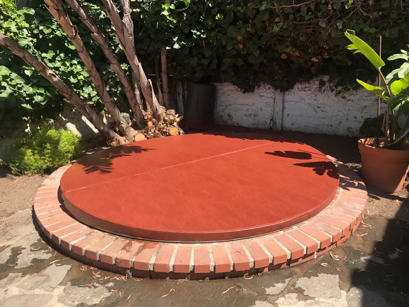 A round rust-brown spa cover on a raised in-ground spa ringed with brick, in a shaded corner garden with a climber-covered wall, a multi-stemmed tree and a large-leaved potted plant.